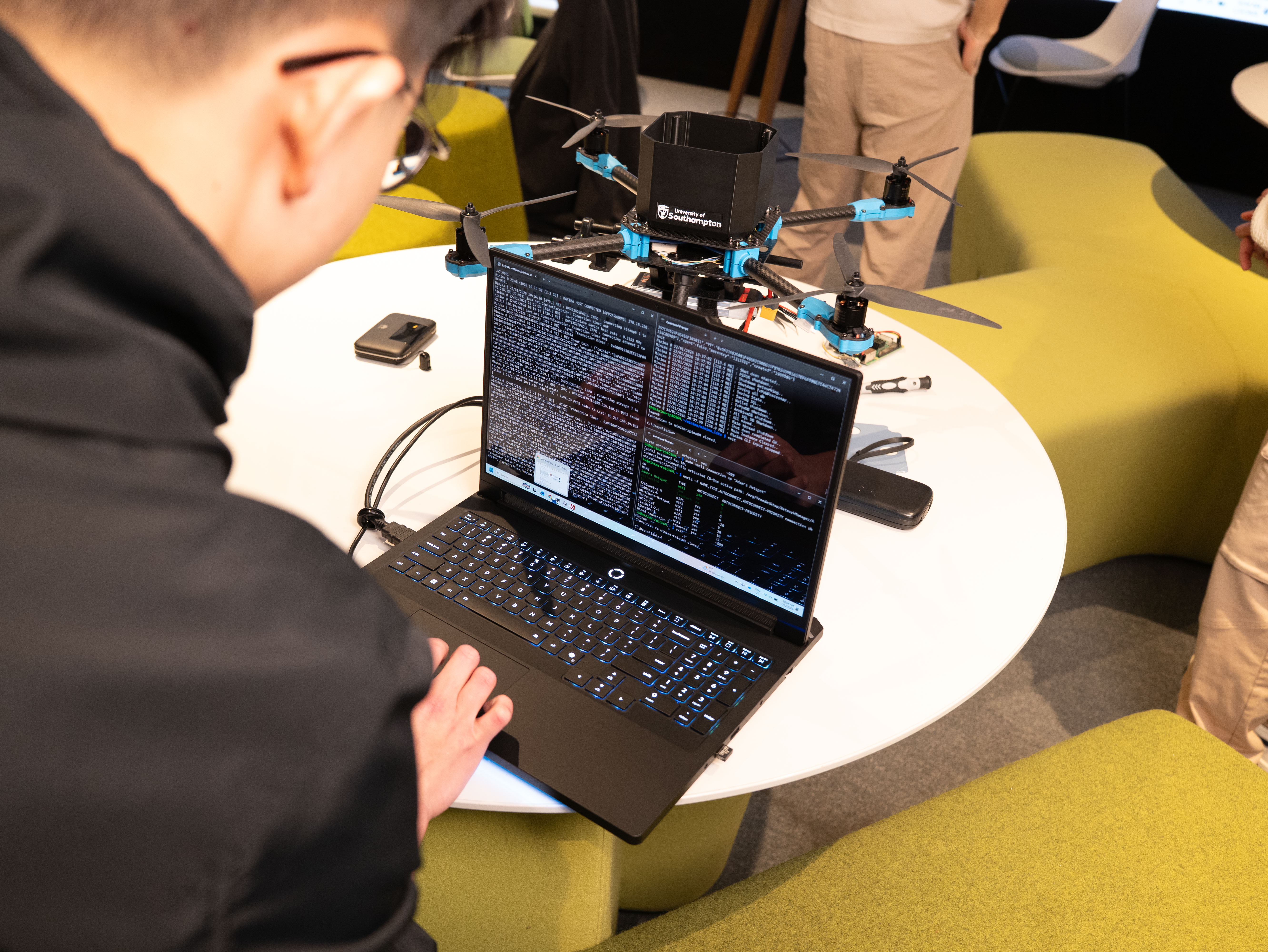 A close-up of a person working on a laptop displaying lines of code, connected to a quadcopter drone resting on a round table. The drone has visible propellers and a central black housing labelled with the University of Southampton logo, with other people standing nearby in a collaborative workspace.