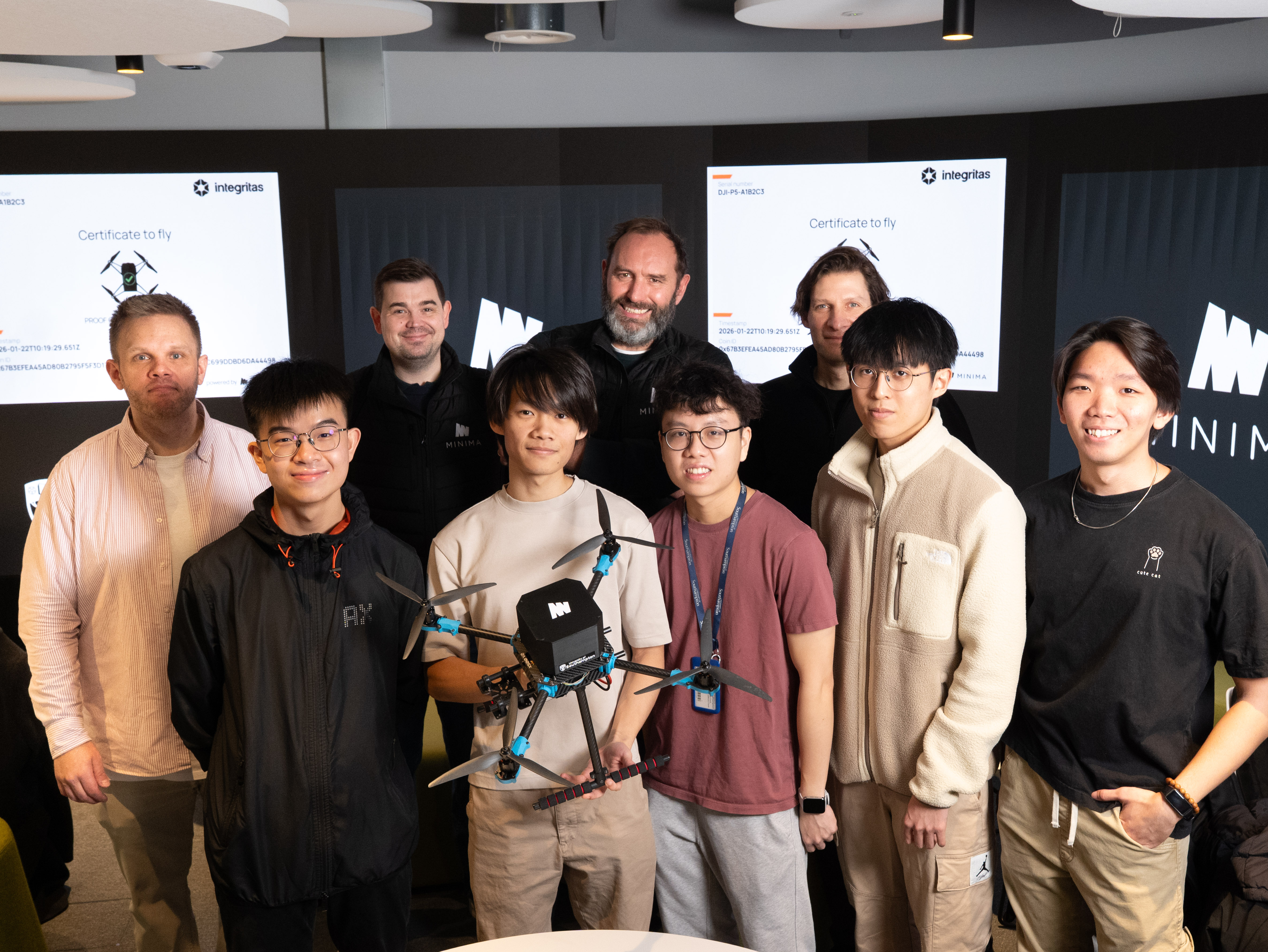 A group of eight men pose indoors with a quadcopter drone, one holding it at the centre. They stand in front of display screens showing a 