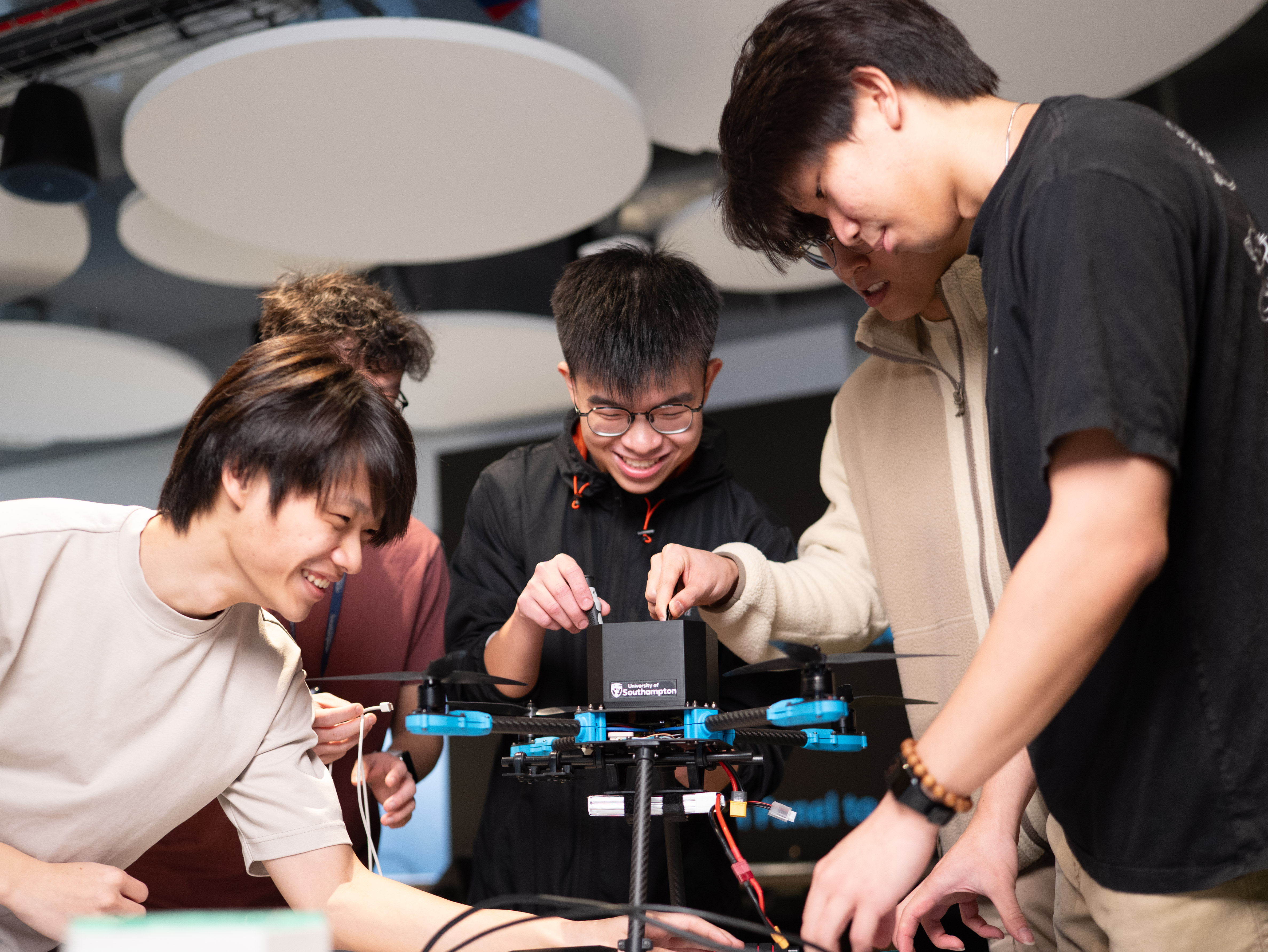 Four university students gathered around a quadcopter drone indoors, smiling as they adjust components on top of it. The drone is mounted on a stand and has blue arms and visible wiring, with a small black box attached to its center. The students appear focused and collaborative, working together in a modern lab or classroom setting with circular ceiling panels overhead.