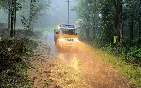 A yellow truck driving along a wet and muddy road with trees and undergrowth either side. It is raining very heavily