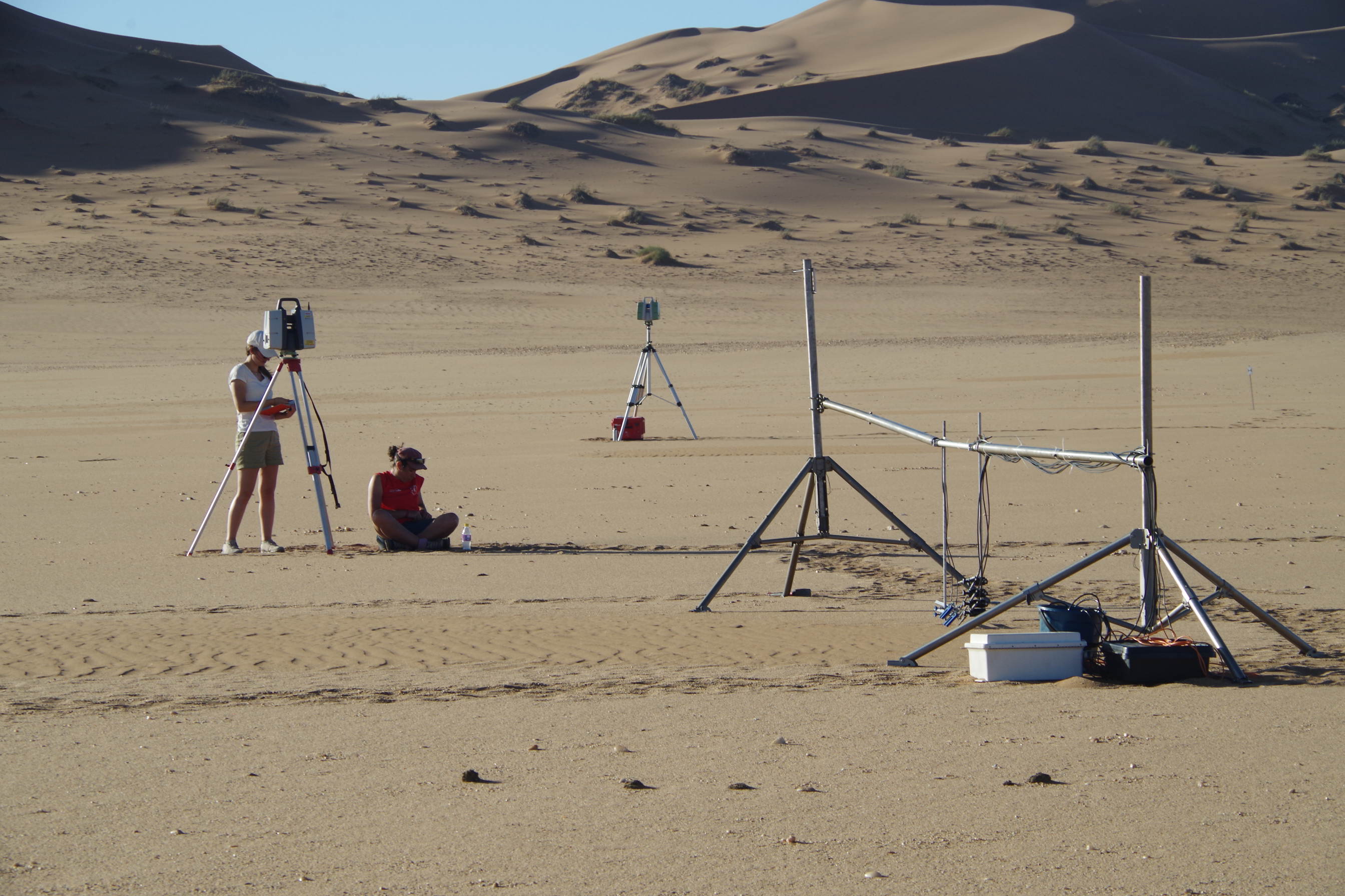 Two people are conducting field research in a desert environment with sand dunes in the background. One person is standing next to a tripod-mounted scanning device, while the other sits on the ground near equipment. Additional scientific instruments on tripods and metal frames with cables are positioned around them