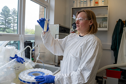 A researcher wearing protective gloves, safety glasses and a lab gown examines a sample tube in a laboratory,