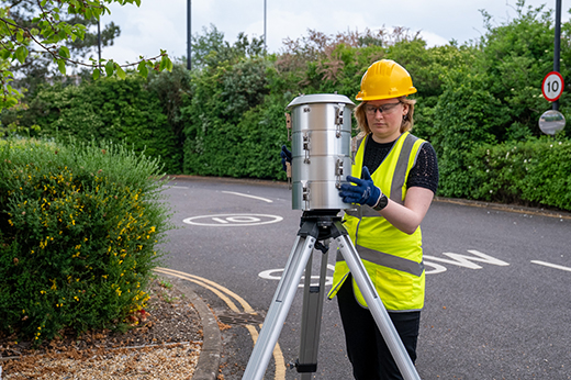 A researcher wearing a hard hat, high-visibility vest and safety glasses adjusts an air monitoring instrument mounted on a tripod beside a road, surrounded by trees and shrubs.