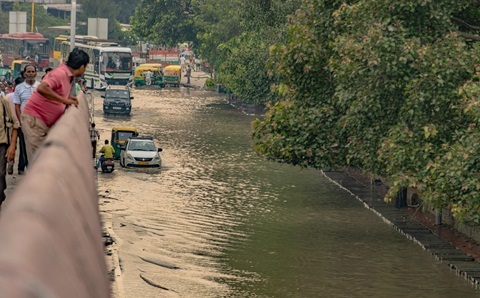 A man in a red T-shirt is leaning over a wall, looking down onto a flooded road below. There is a car, a motorbike and a tuk-tuk driving through the water, with a van and a bus a bit further behind. The other side of the road is lined with green trees.