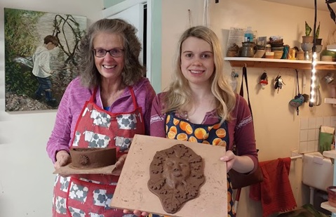 Two women standing in an art studio holding items made from clay