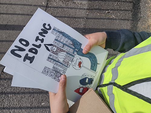 A child wearing a high-visibility vest holds illustrated anti-idling leaflets beside a road. One leaflet reads 