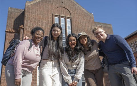 Five people standing close together with arms round each other’s shoulders, smiling. They are outside, in front of a large red brick building, and under a clear blue sky. 