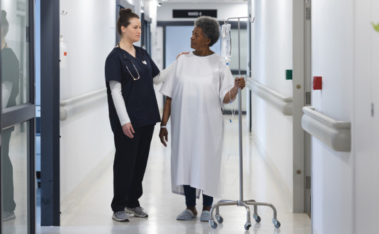 A nurse dressed in dark blue uniform standing next to a patient in a white gown holding a wheeled drip stand in a hospital corridor.