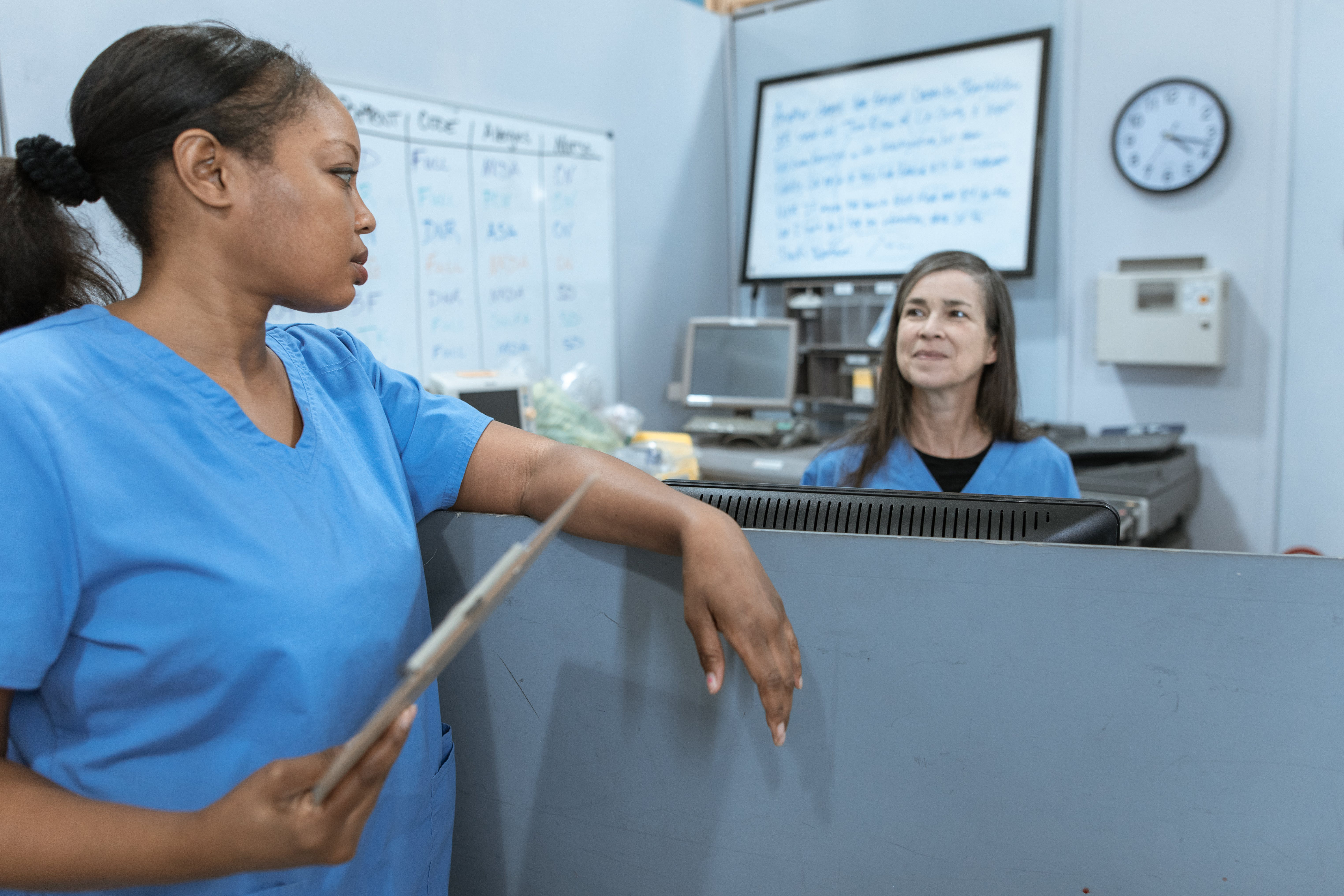 Two nurses wearing blue uniforms talking to each other across a ward reception desk.