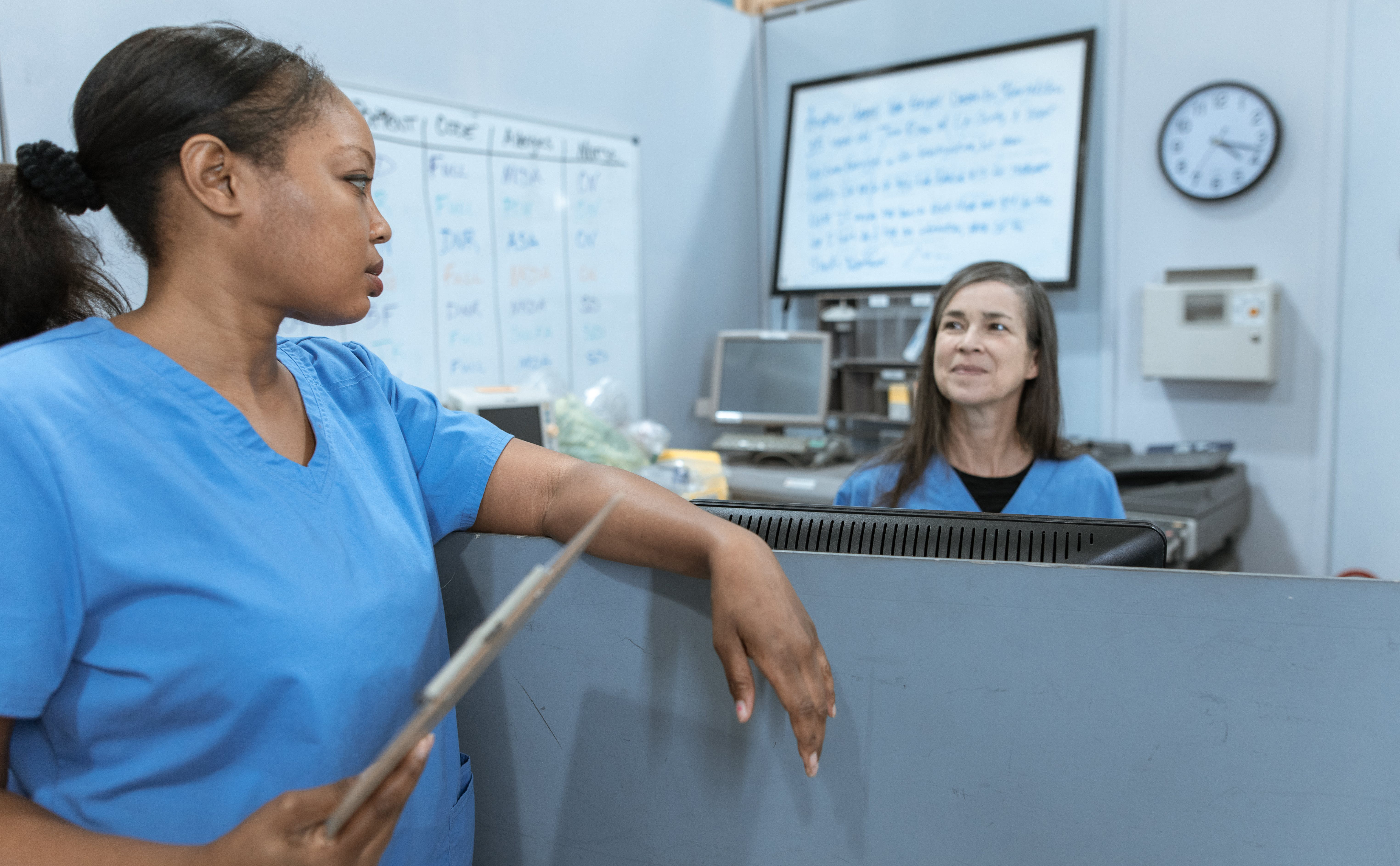 Two nurses wearing blue uniforms talking to each other across a ward reception desk.