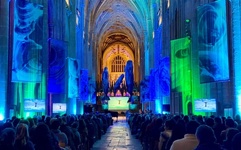 A large audience sat inside a cathedral, with blue and orange lighting. There are banners featuring marble-like art either side, and sculptures of 3 whales hanging in the distance of the image.