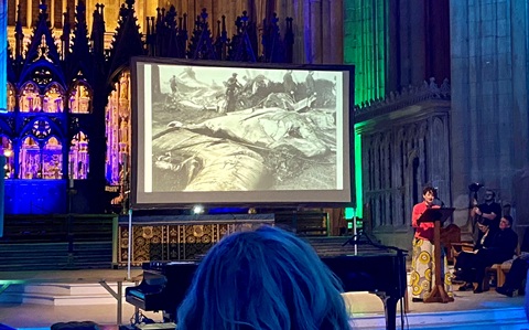 A woman in a red top and long patterned skirt standing at a lecturn inside a cathedral. Behind her is a screen showing a black and white image of a whale carcass.