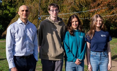 Four people stood in a row, facing the camera and smiling, they are stood outside in front of greenery