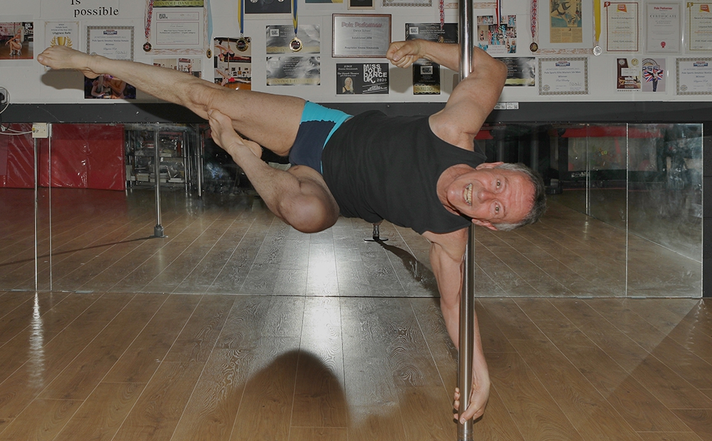 A man wearing a black vest and blue shorts, holding himself up horizontally from a silver pole