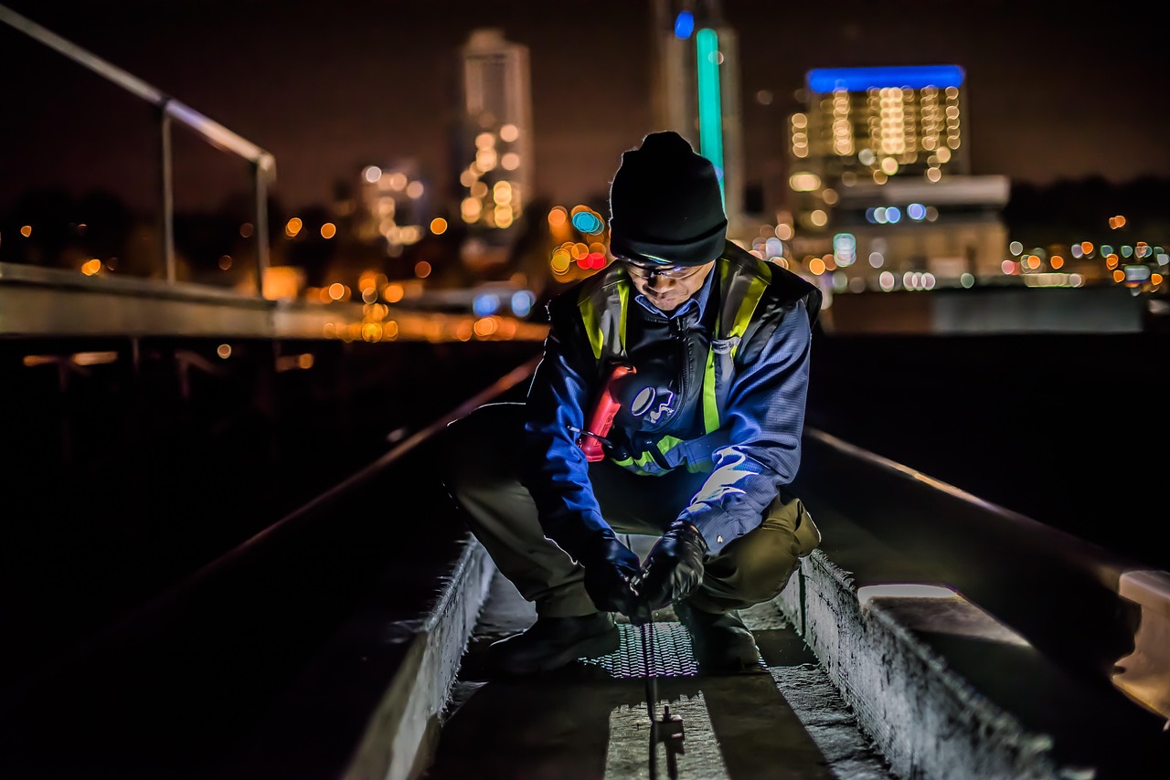 A construction worker wearing a safety vest, gloves, and a beanie is kneeling on a railway track at night, illuminated by a headlamp. The background features a city skyline with colorful, out-of-focus lights.
