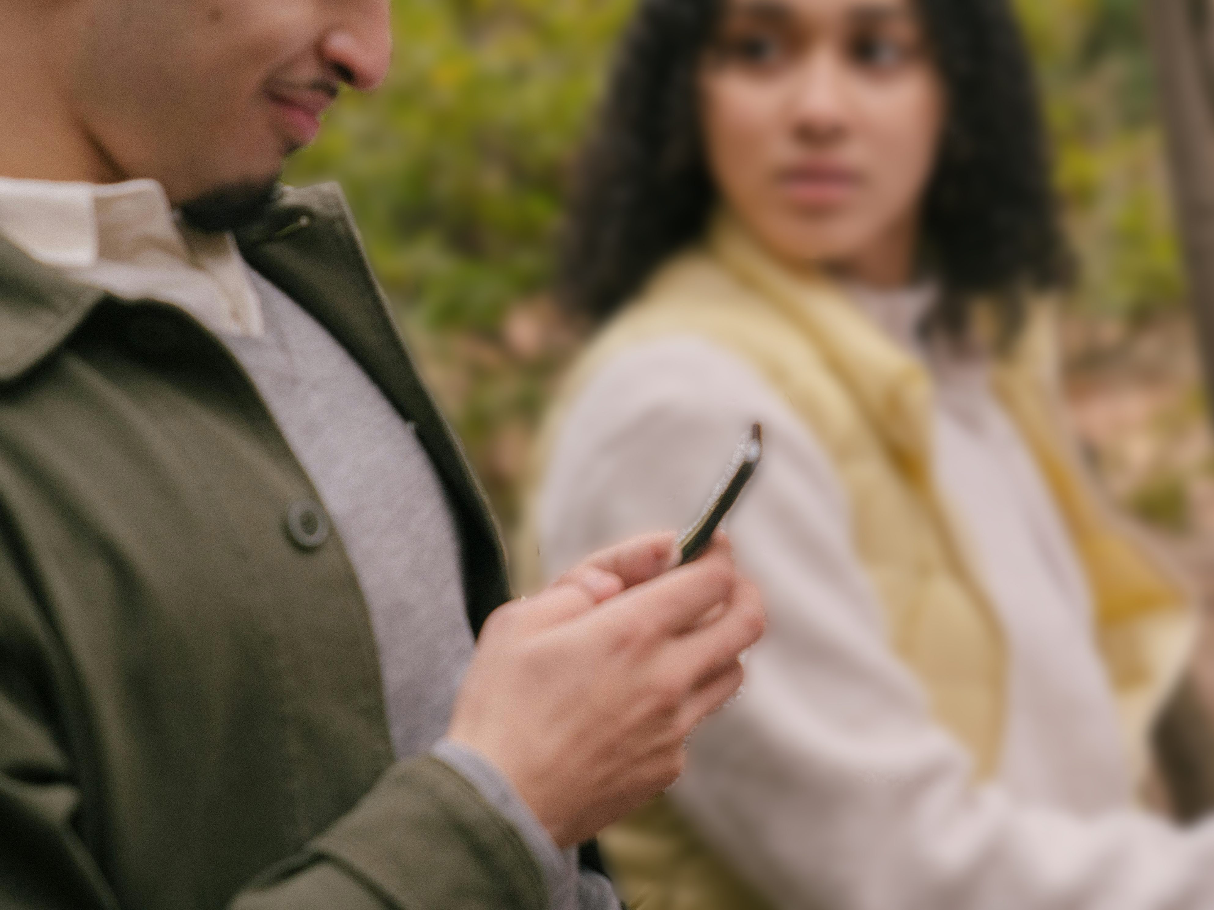 A man looks down at his phone while sitting next to a woman, who looks at him with a displeased or concerned expression. The scene suggests a moment of distraction or tension between them.