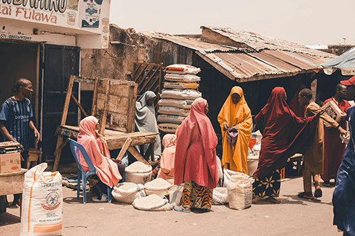 People browse and buy sacks of rice and other staple foods at a busy open-air market. Several women wearing brightly coloured hijabs stand around grain-filled bags outside weathered market stalls with corrugated metal roofs, while other shoppers and vendors move through the scene on a hot, sunny day.