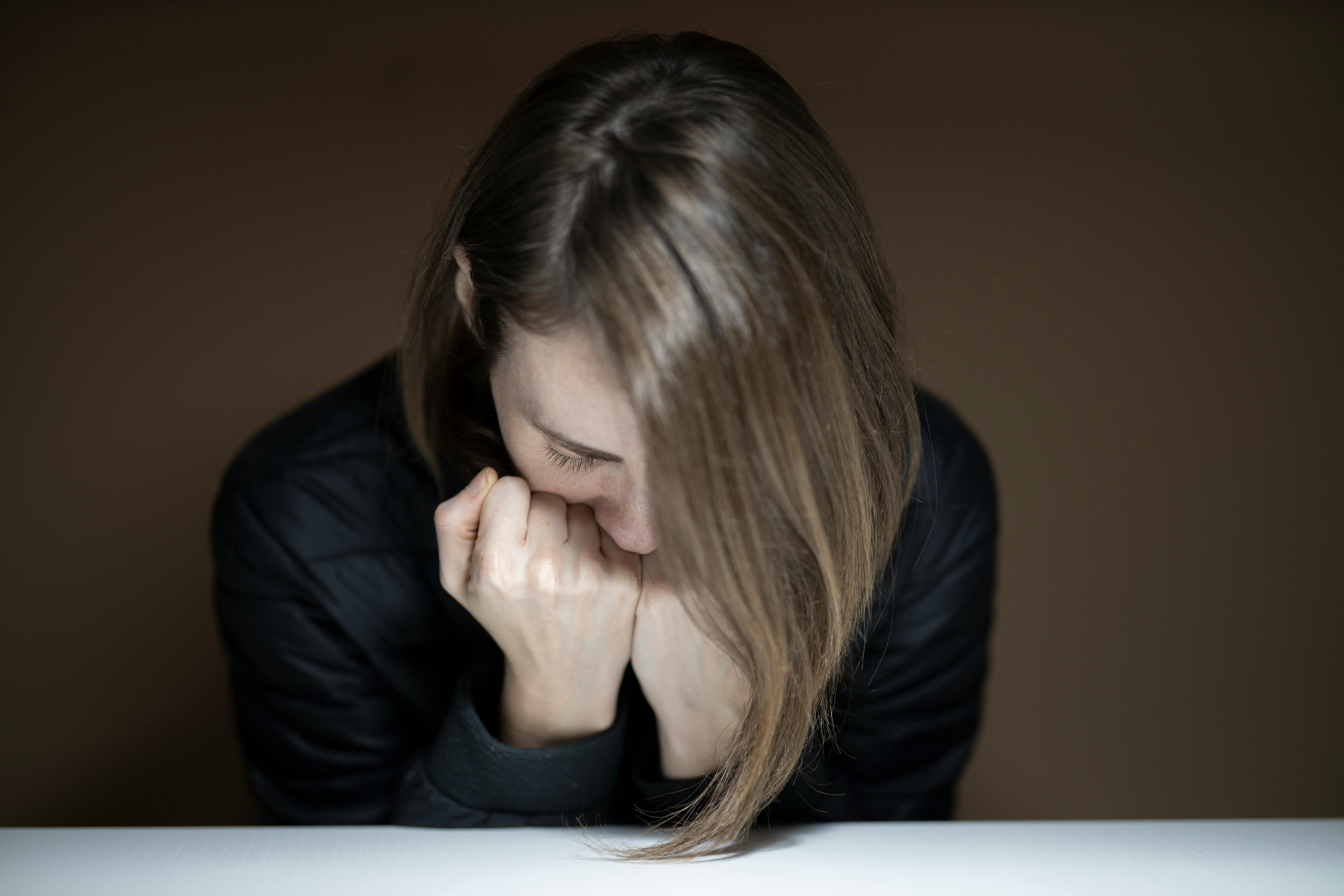 A woman with long brown hair sitting with her head bowed and hands pressed against her face, appearing distressed or deep in thought, against a dark background.