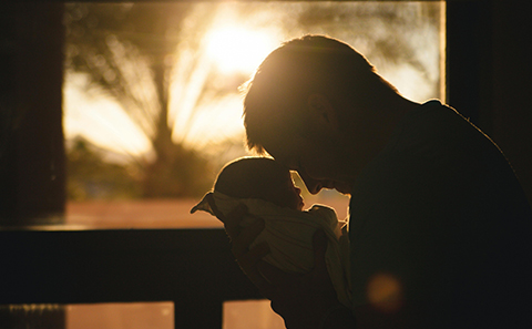 A silhouetted man gently holds a newborn baby up to their face near a window, with warm sunlight glowing behind them.