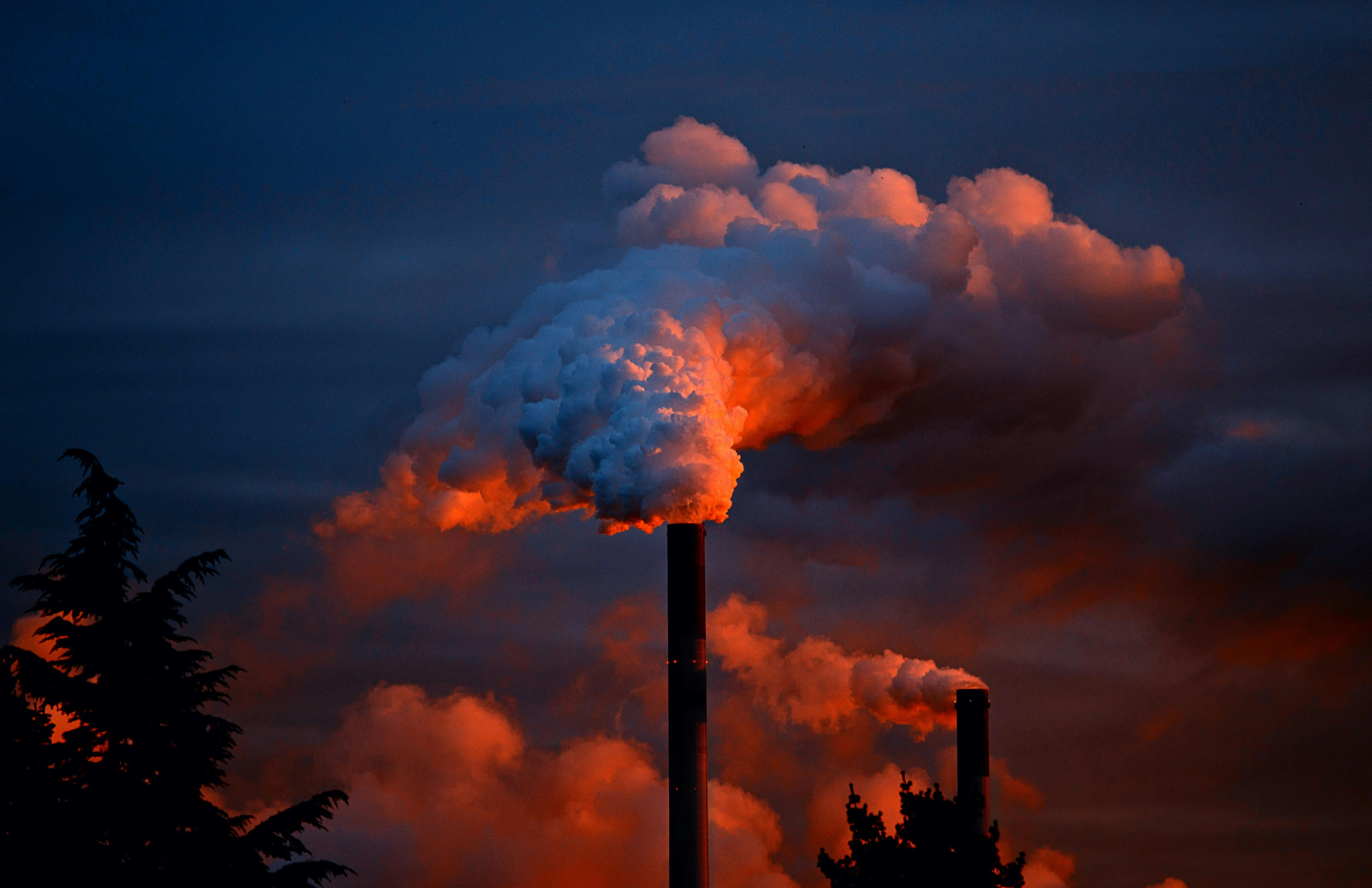 ’Two large smokestacks emit thick plumes of smoke, illuminated in shades of red and orange by the sunset or artificial lighting, contrasting against a dark blue evening sky. Silhouetted trees appear in the foreground, adding depth to the image and emphasizing the industrial scene.’