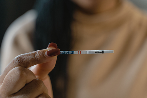 Close-up of a person holding a pregnancy test strip between their fingers, with two visible lines on the test indicating a positive result. The background is softly blurred, showing part of the person's face and beige clothing.