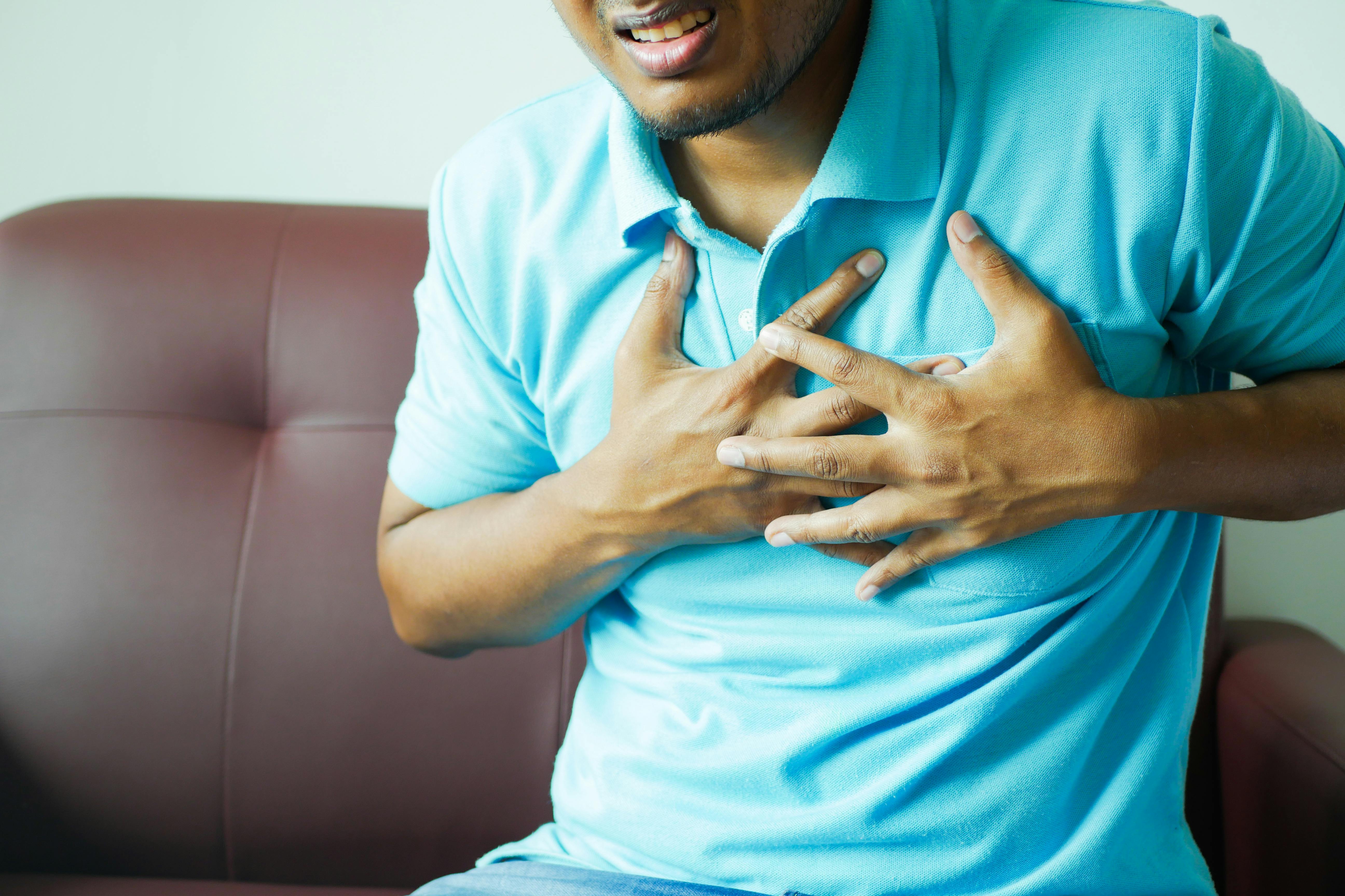 A man wearing a light blue polo shirt is clutching his chest with both hands, appearing to be in distress or pain, possibly experiencing chest discomfort. He is sitting on a brown couch with a tense expression on his face.