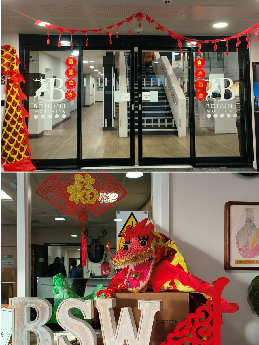 A school entrance is decorated with red lanterns, hanging ornaments, and a decorative dragon for a Lunar New Year celebration. Inside, a display features a bright dragon head and large letters spelling “BSW.”