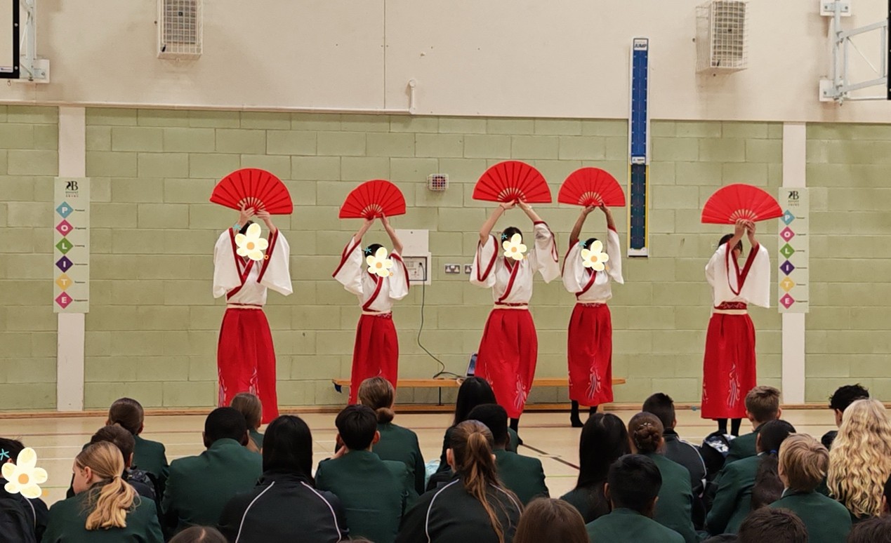A group of performers in red skirts and white tops hold large red fans above their heads while dancing in a school gym. Students seated on the floor watch the performance.