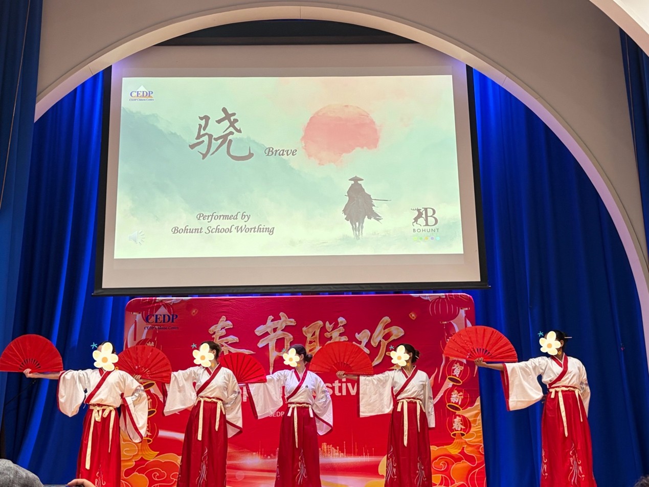 Performers dressed in red and white traditional-style outfits hold red fans on a stage beneath a large screen reading “Brave,” with festival decorations behind them. The scene is part of a cultural celebration in an auditorium.