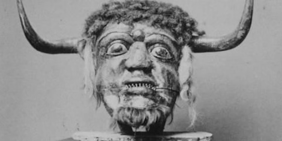 This is a black and white photograph of what appears to be an ancient carved mask or sculpture with exaggerated facial features including bulging eyes, bared teeth, and curly hair or texture on top. The figure has two horn-like protrusions extending from either side of its head, giving it a demonic or mythological appearance.