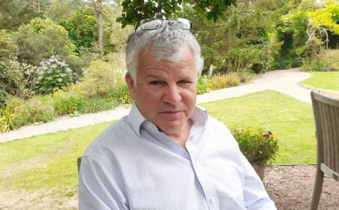 A grey-haired man wearing a collared shirt and shown seated outdoors