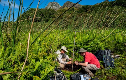 Two men crouching in a lush green wild area, facing one another. There are mountains in the background and the sky is deep blue.