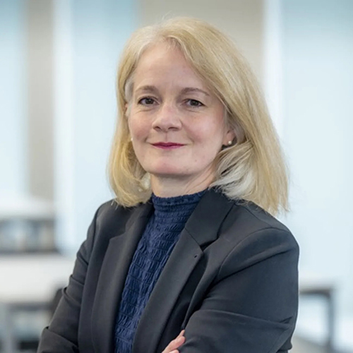 Professional head-and-shoulders portrait of a woman with shoulder-length blonde hair, wearing a dark blazer over a navy high-neck top. She is seated with her arms folded, looking directly at the camera with a slight smile. The background is a softly blurred office or institutional setting with large windows and neutral tones.