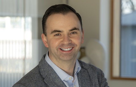Head and shoulders photo of a man with dark hair wearing a grey suit, smiling