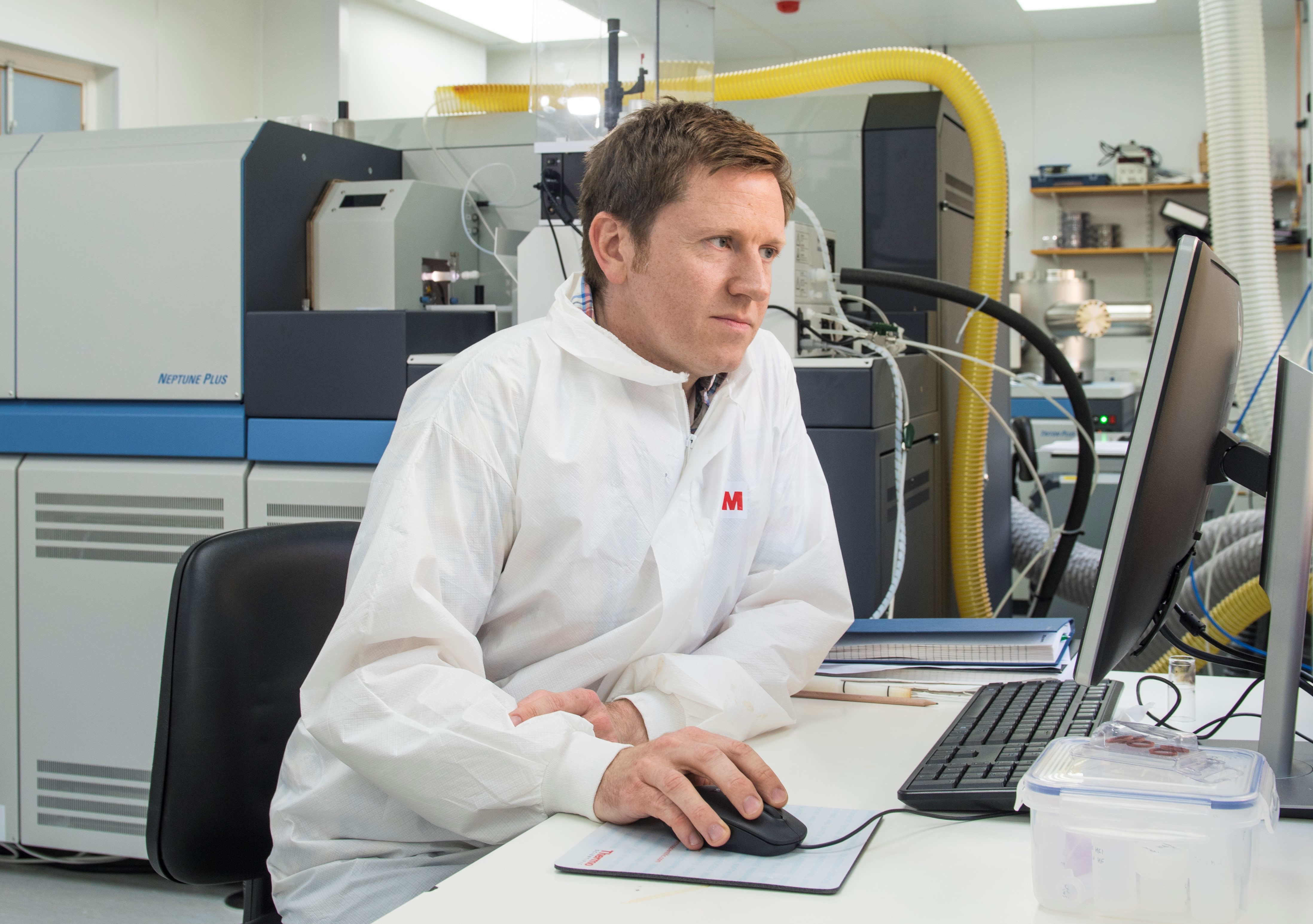 Man in white coat sitting at a desk surrounded by lab equipment.