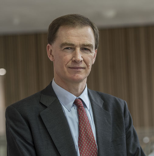 Head and shoulders view of a man wearing a dark grey suit jacket, light shirt and red patterned tie.
