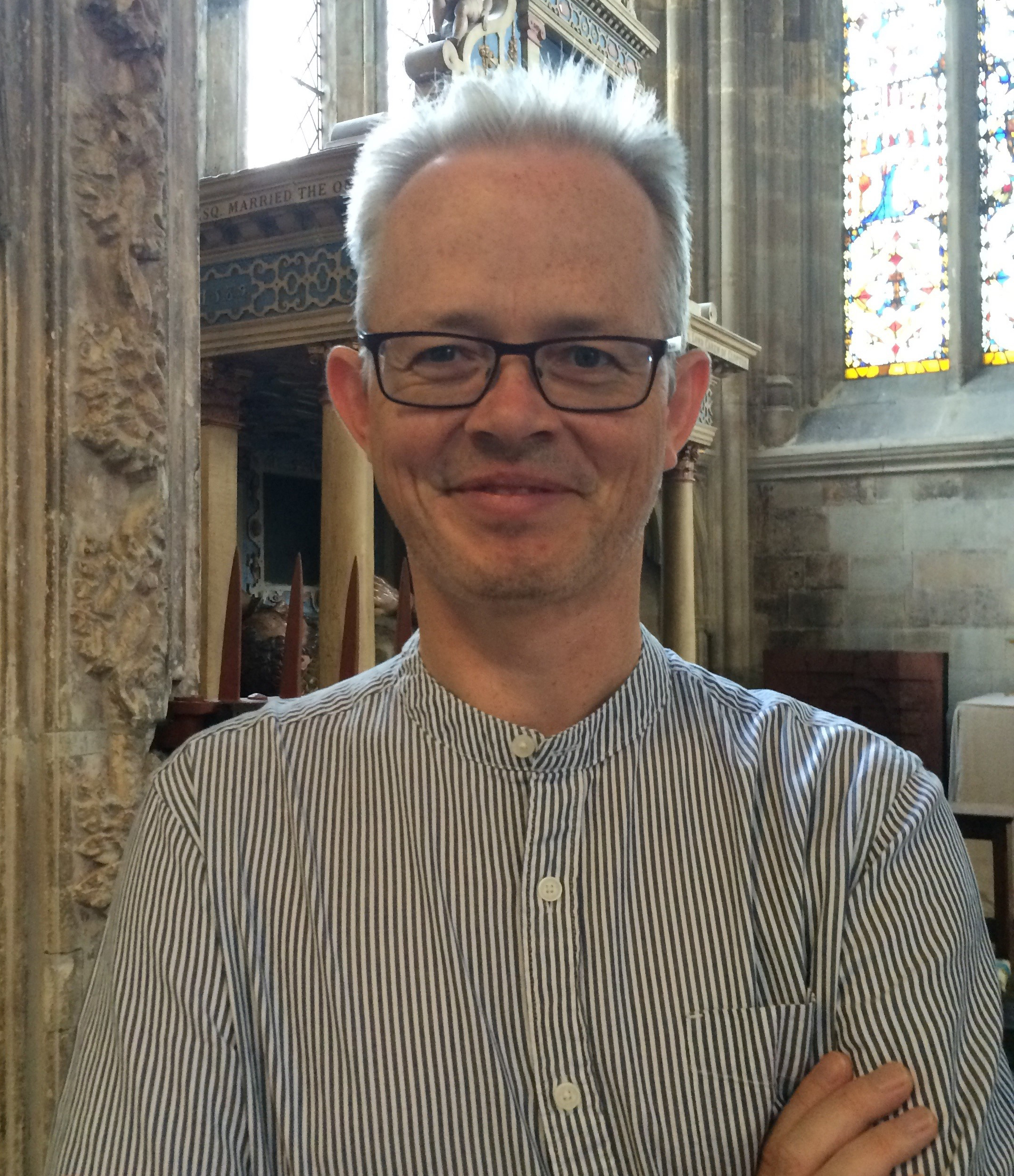 A head and shoulders view of man wearing a striped collarless shirt and glasses, stood with arms crossed in a church.