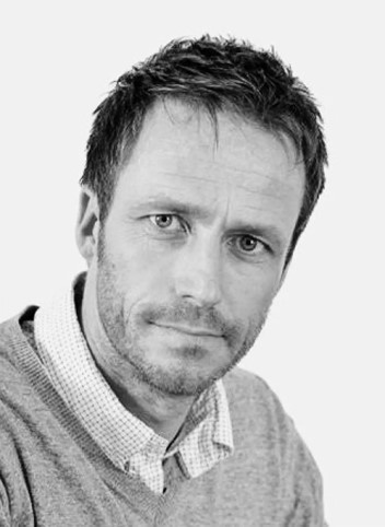 Black and white image of a man in head and shoulders view, wearing a checked shirt and v-neck jumper, looking to the camera.