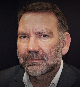 Head and shoulders image of a man with a beard, wearing an open neck white shirt and dark jacket.
