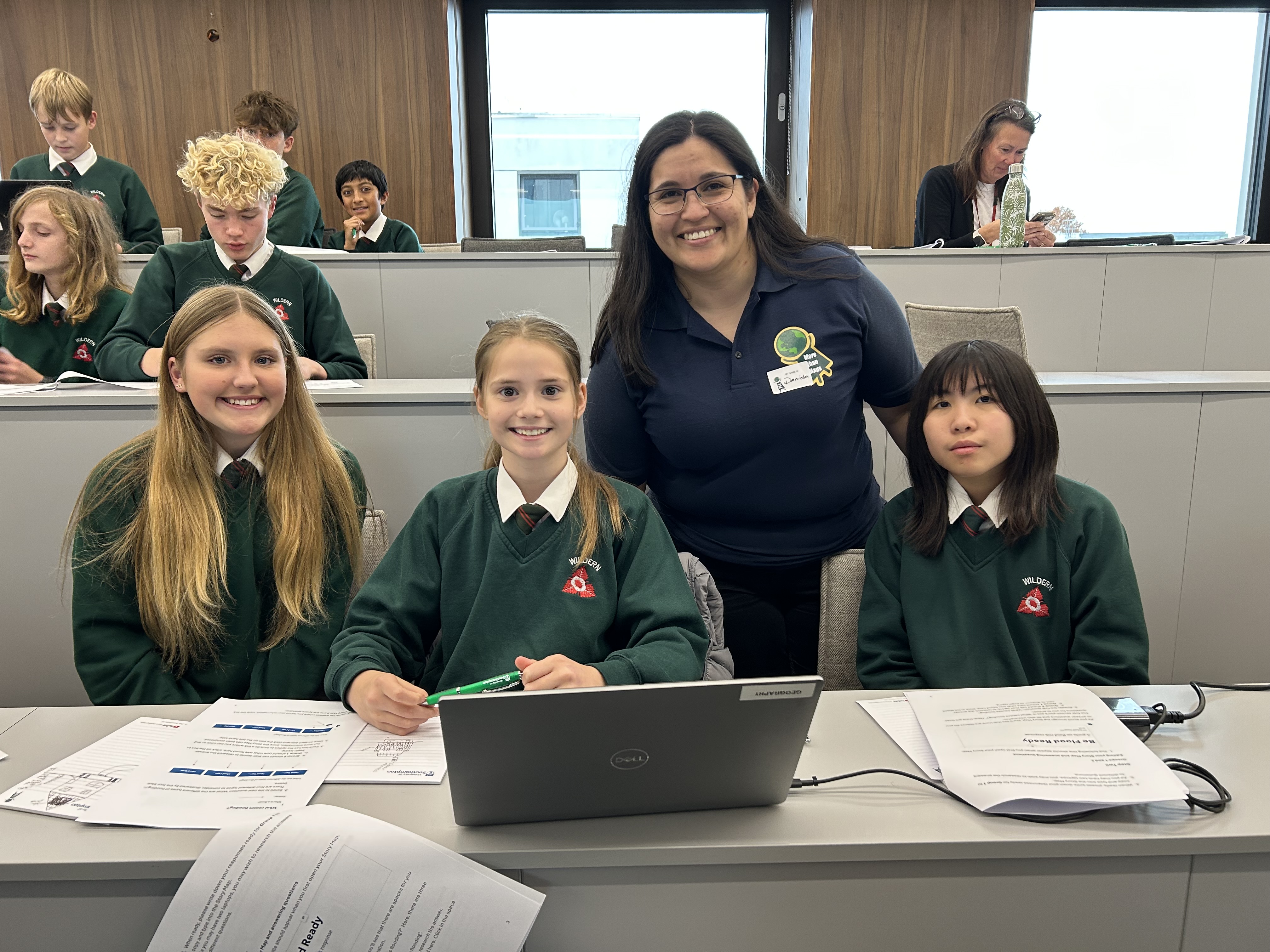 A classroom with students in green sweaters and a teacher. They are seated in front of a laptop and papers.