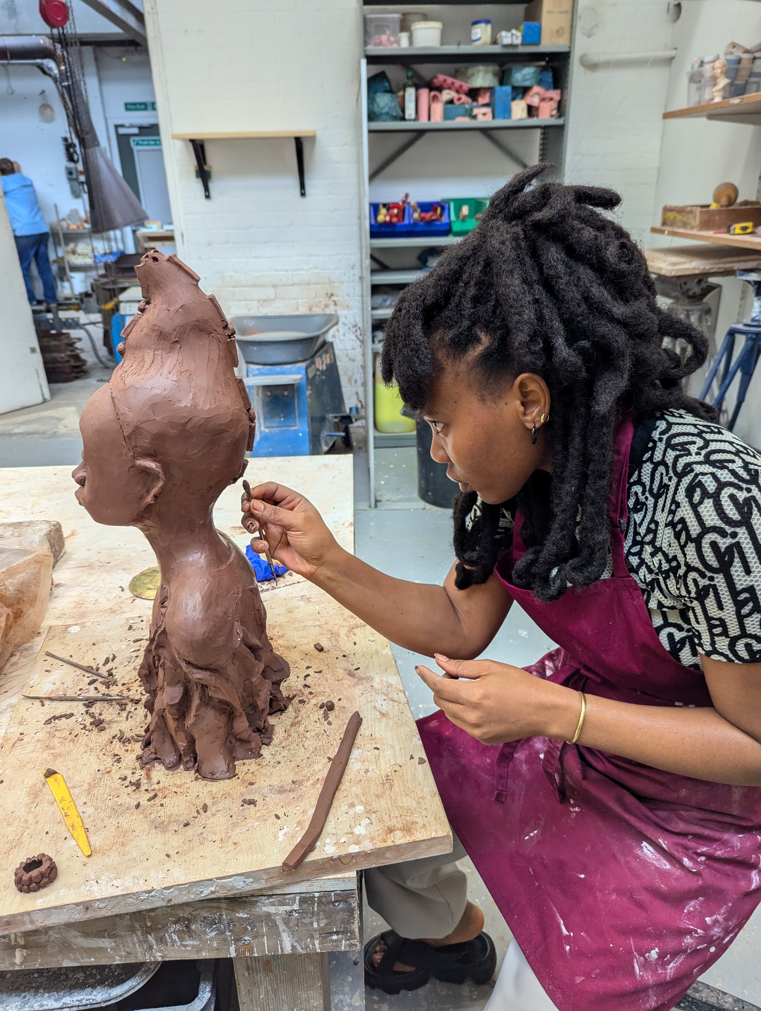 A person wearing a maroon apron is shaping a tall clay sculpture with organic, root-like details at the base on a wooden worktable. The workspace includes sculpting tools, clay pieces, and shelves filled with pottery materials in the background.