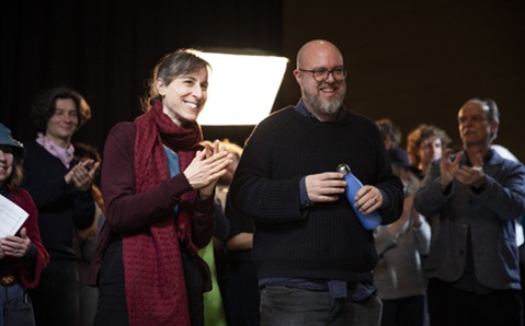 A woman with dark hair tied up wearing a dark red jumper and long scarf, stood next to a man with a beard wearing glasses and a dark jumper. They are stood inside, and out of focus behind them is a group of people clapping
