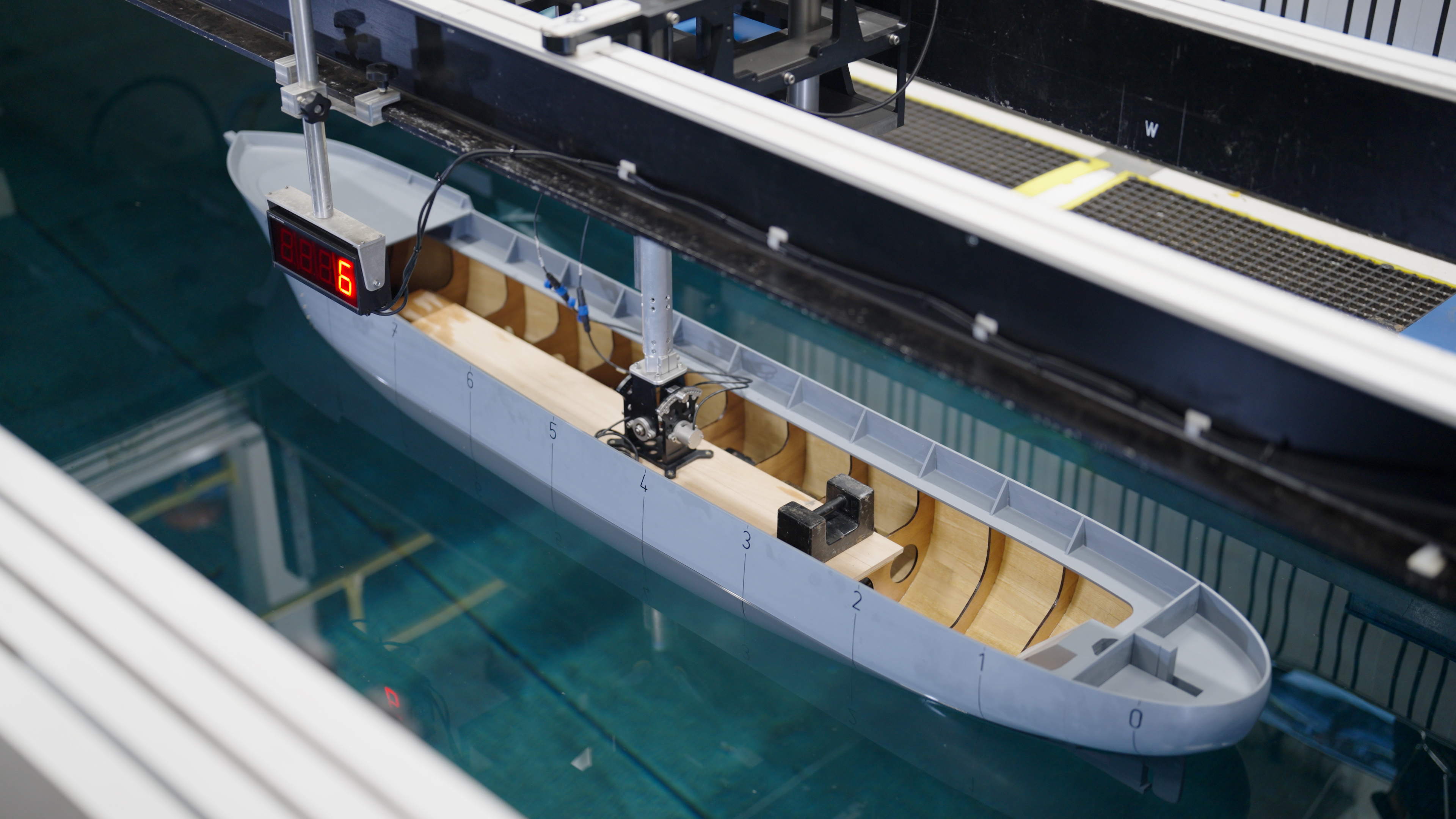 A model ship hull being tested in a towing tank. The grey and wooden model is partially submerged in clear blue water, attached to metal measuring equipment and sensors. A digital display with the number 6 is visible, indicating test data being recorded.
