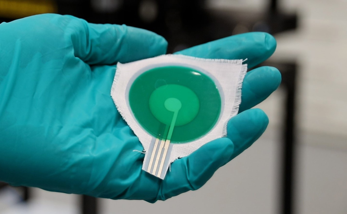 Round green sensor embedded in swatch of material being held in the palm of a hand wearing a green surgical glove
