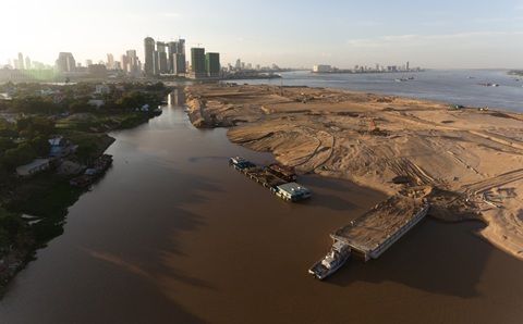 Aerial view of vessels on a river next to a large sand bank. There are high rise buildings of a city in the background