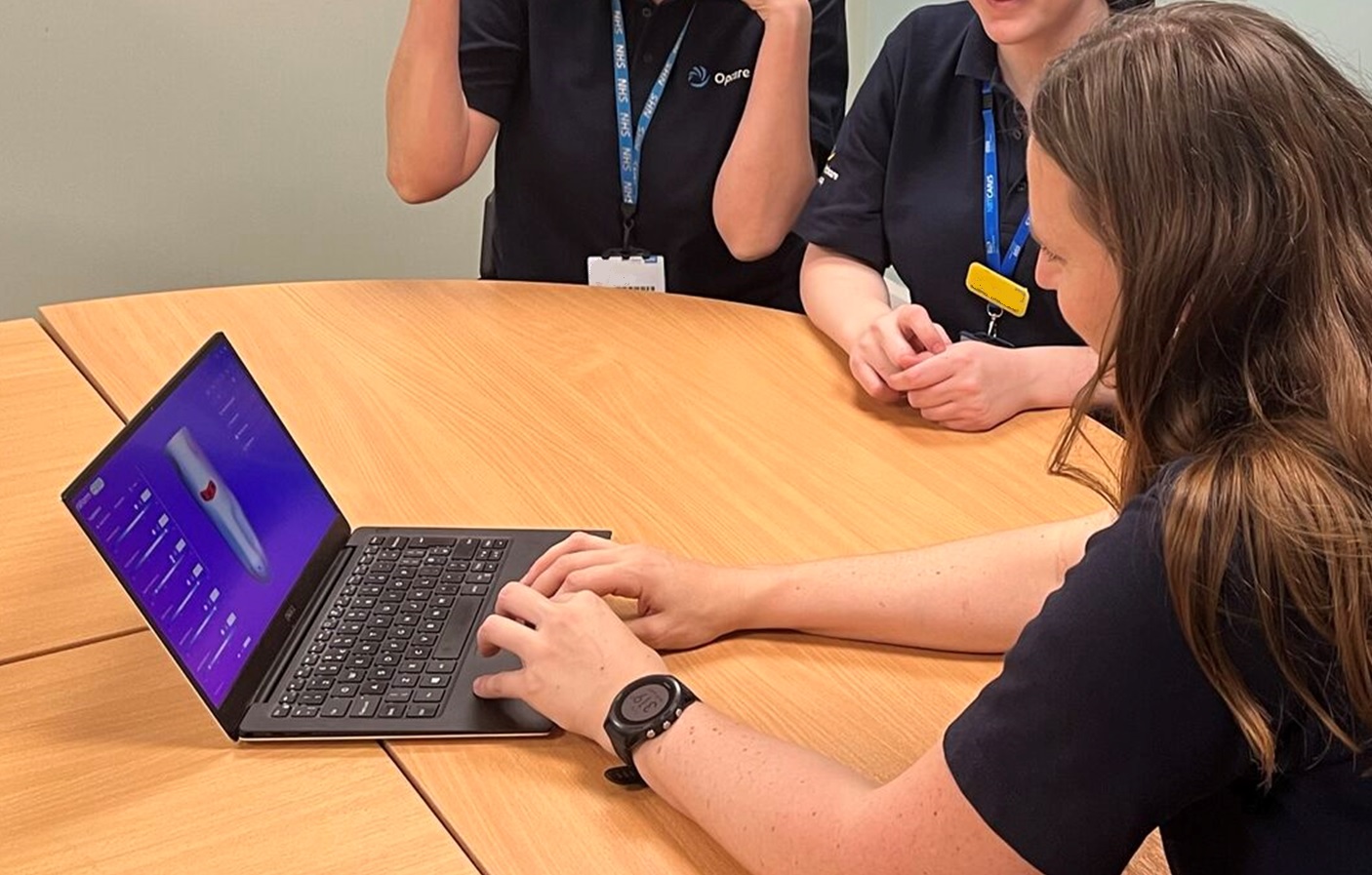 Three people sit around a wooden table. One person is using a laptop that displays a 3D model of a prosthetic socket on the screen. The others, wearing navy uniforms with NHS lanyards, appear to be discussing or observing.