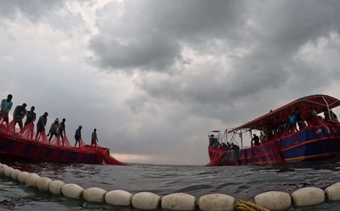 Two fishing boats in the sea. On each boat, a group of people has hold of the edge of a red net, which they are pulling in. There is a row of white floats in the foreground. The skies overhead are cloudy and grey.