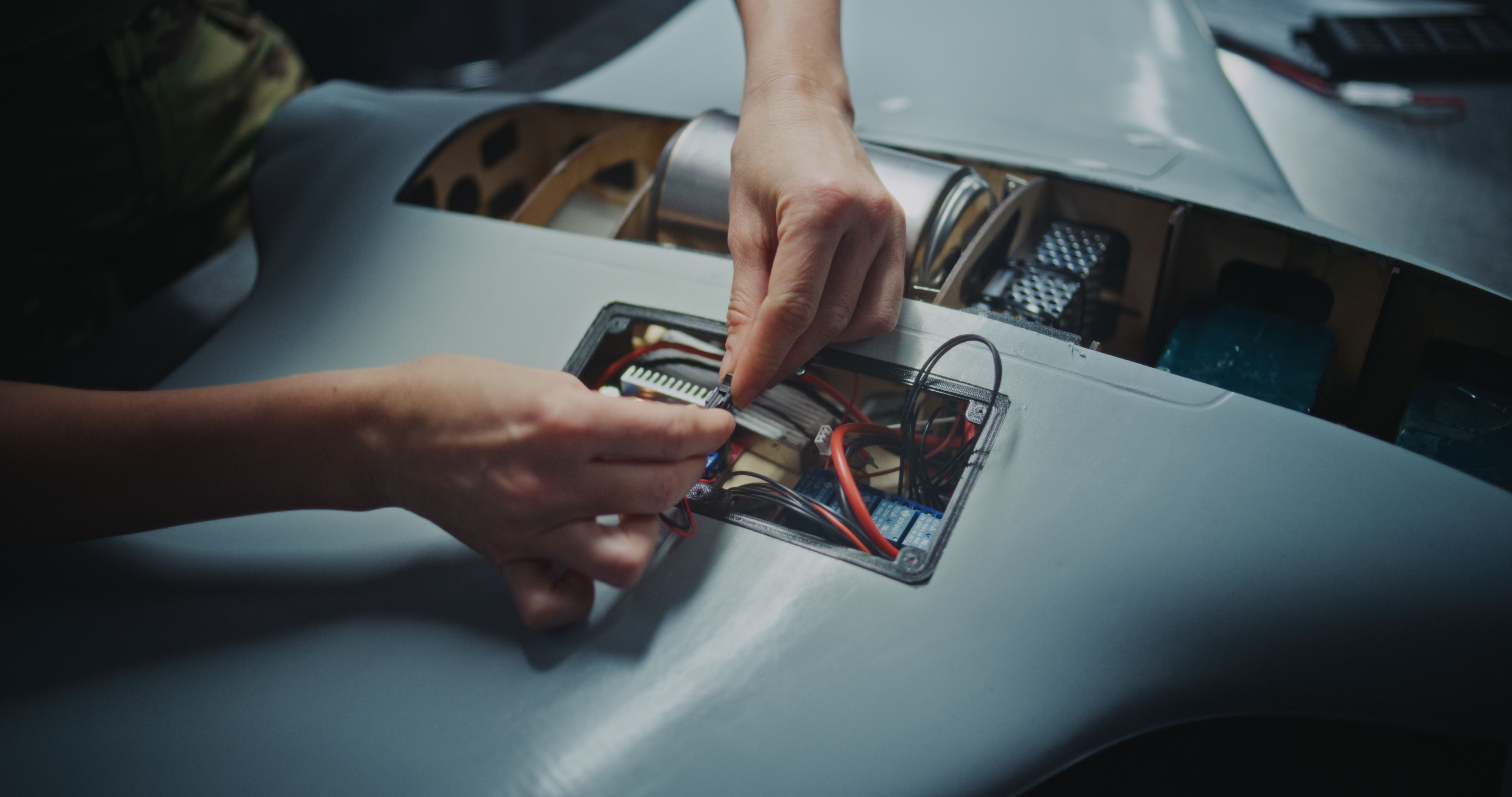 Close-up of hands connecting wiring inside an open panel of a small robotic device, with visible circuit boards, cables and mechanical components