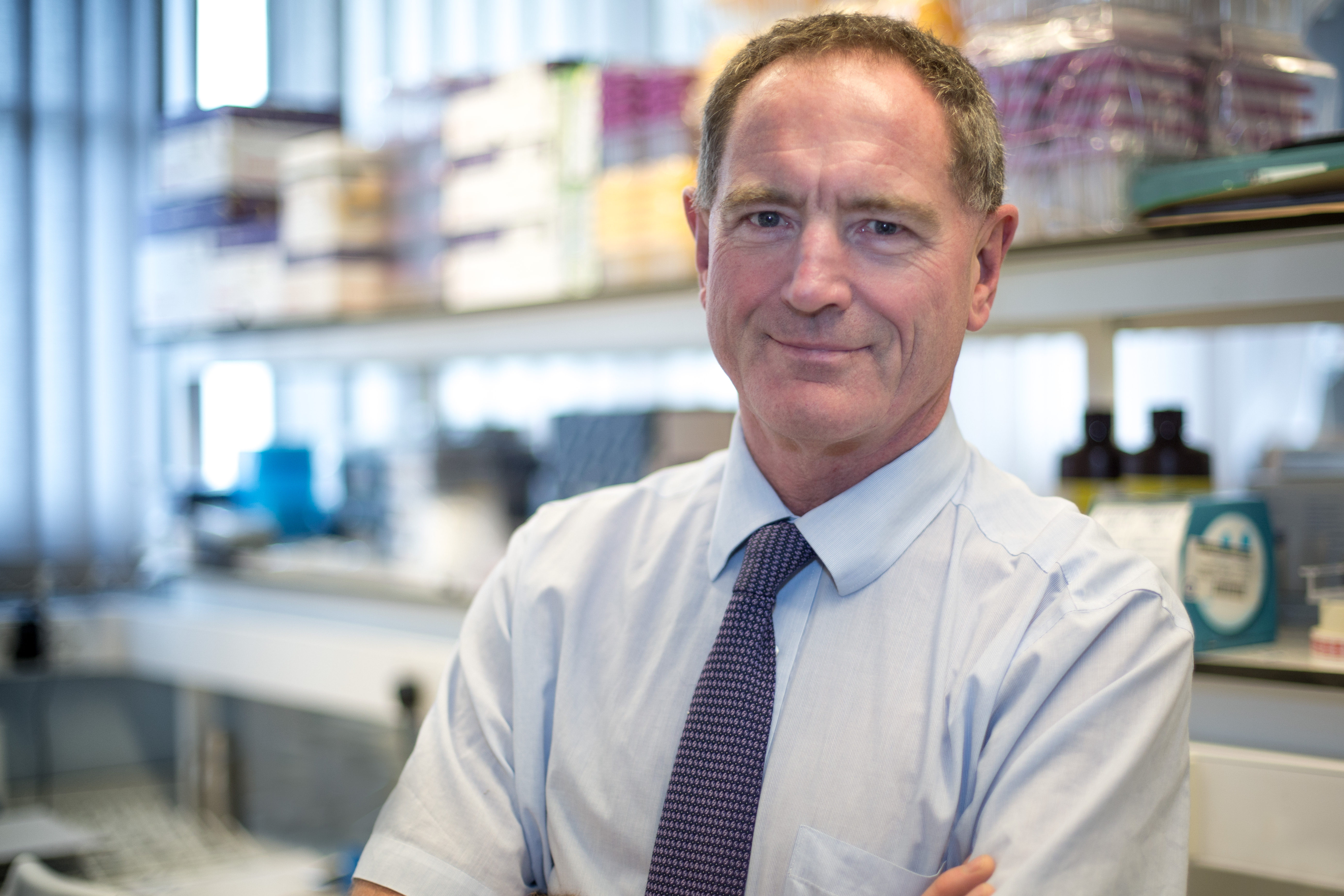 A man with his arms folded wearing a blue tie is looking into the camera with out-of-focus lab shelving behind. 