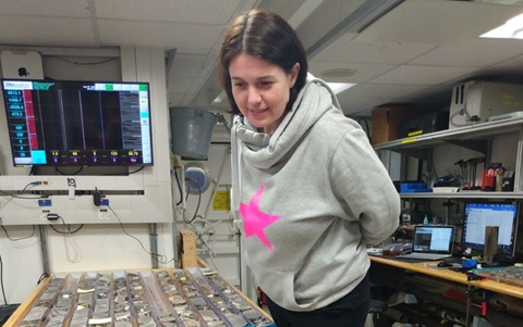 Woman with dark straight hair wearing a grey hoodie with a pink star on the front, standing over a desk with lots of rock sample on it